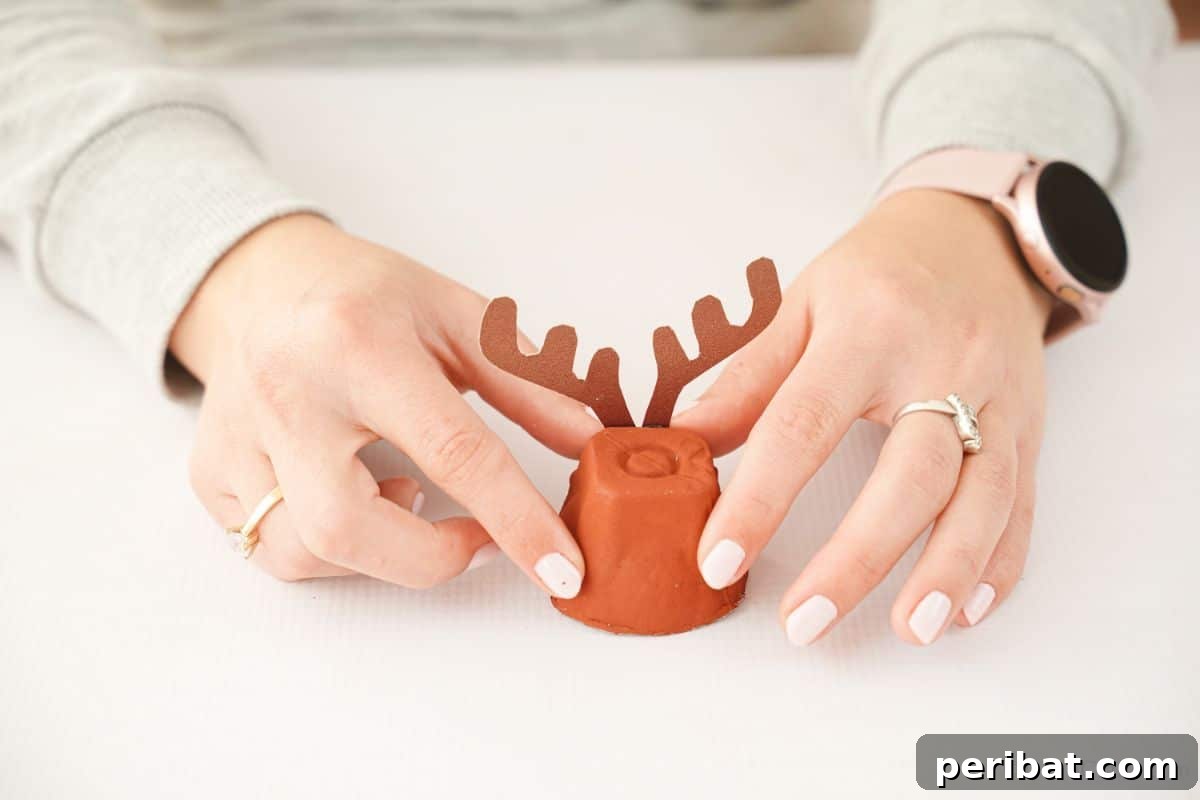 A hand carefully holding cut antlers onto the back of an egg carton reindeer body, demonstrating the assembly step.