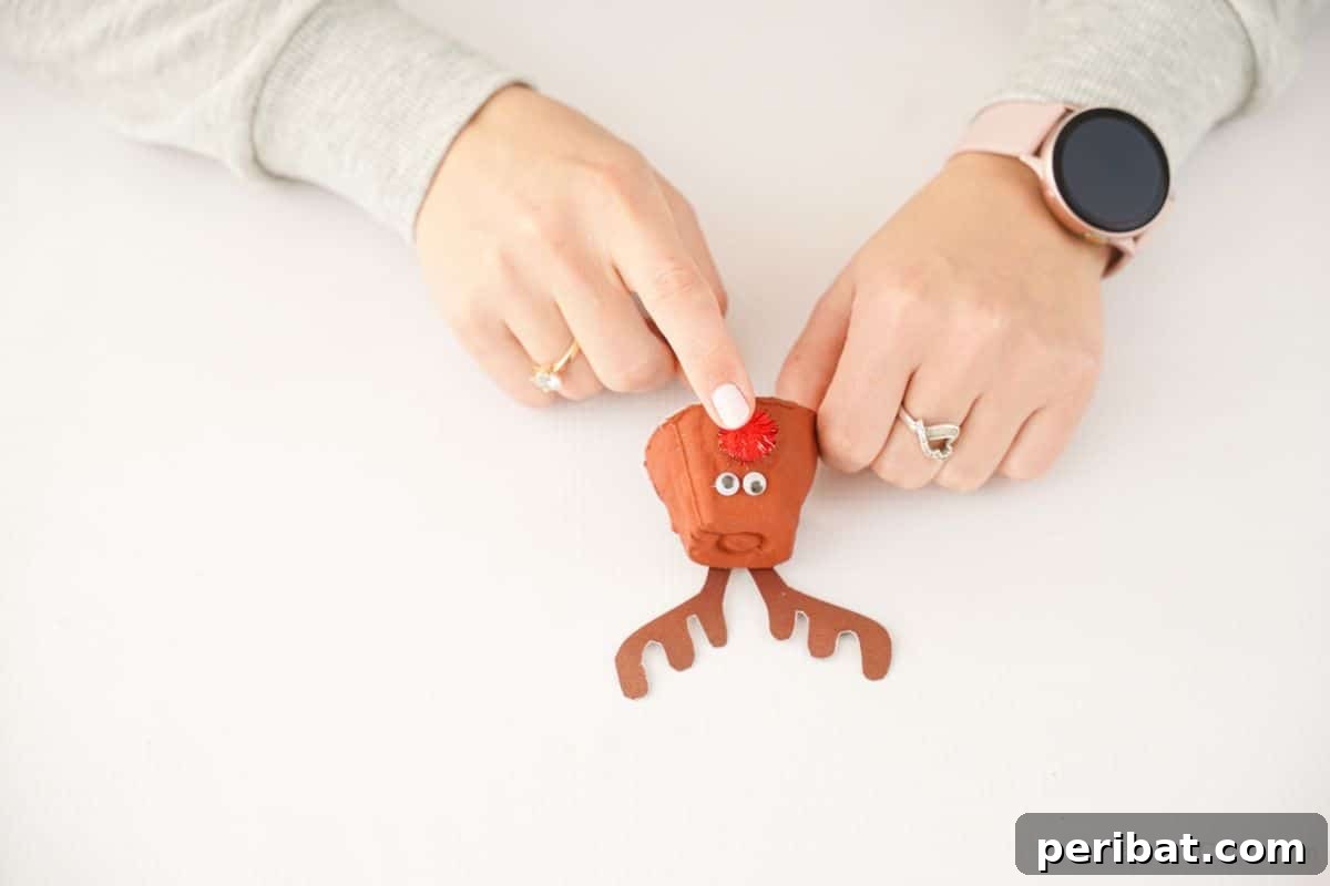 A finger gently holding a red pom-pom onto the front of an egg carton cup, forming the cheerful nose of the reindeer craft.
