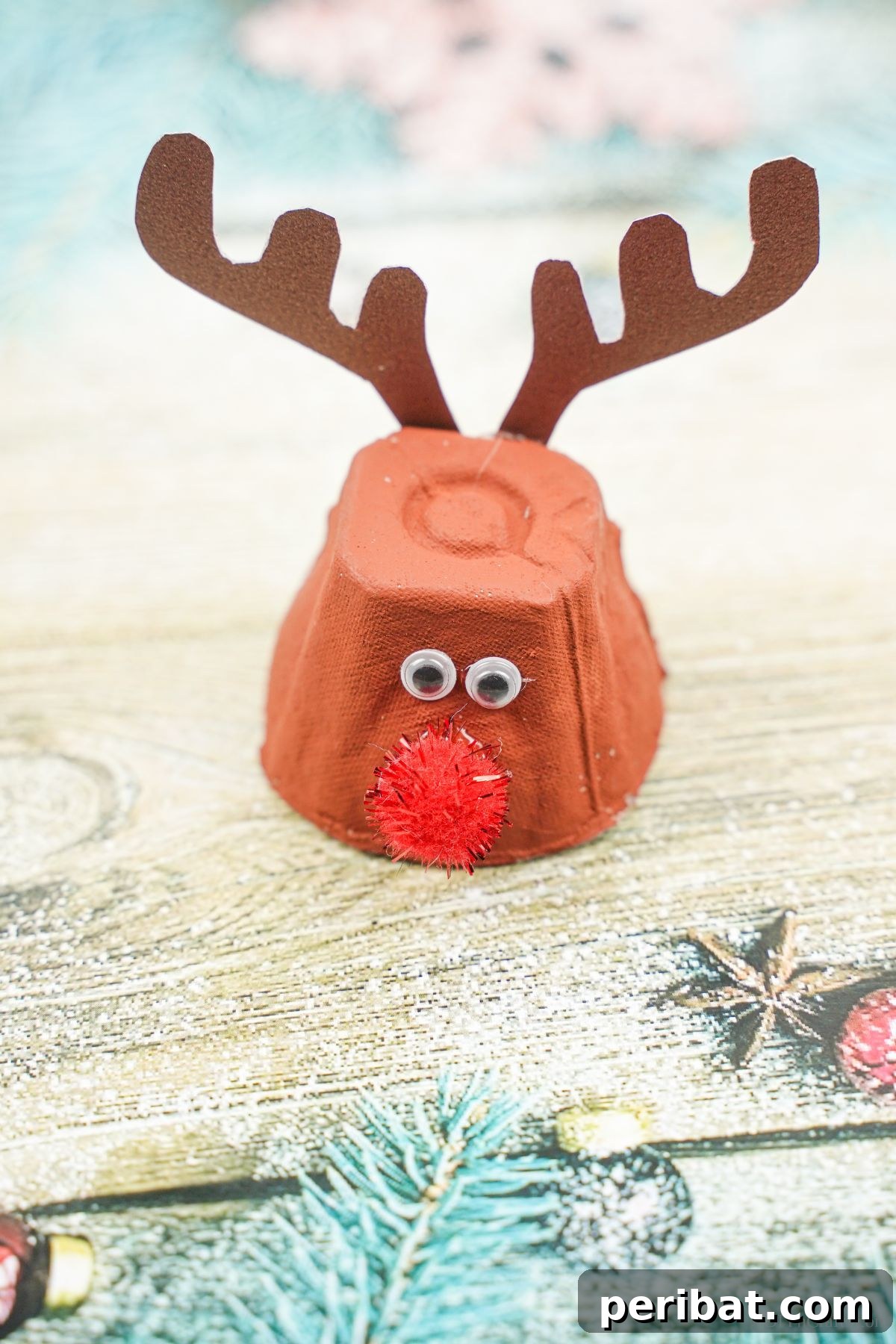 A charming brown egg carton reindeer with a bright red nose, set against a backdrop of blue wood paper, creating a rustic yet festive scene.