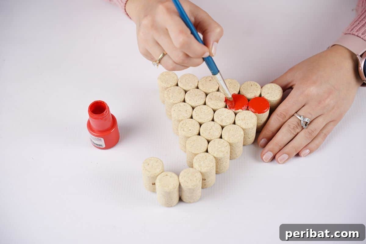 A hand expertly paints a wine cork red, transforming it into a component of a festive Santa hat decoration.