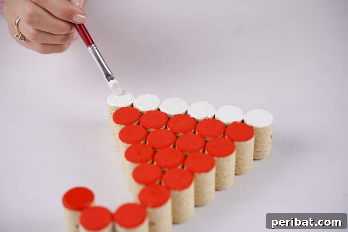 A hand uses a paintbrush to apply white paint to the bottom row of corks, forming the trim of a Santa hat decoration.
