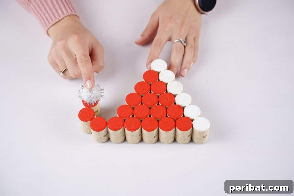 A hand gently holds a sparkly pom-pom onto the curved end of a cork hat, completing the festive decoration.