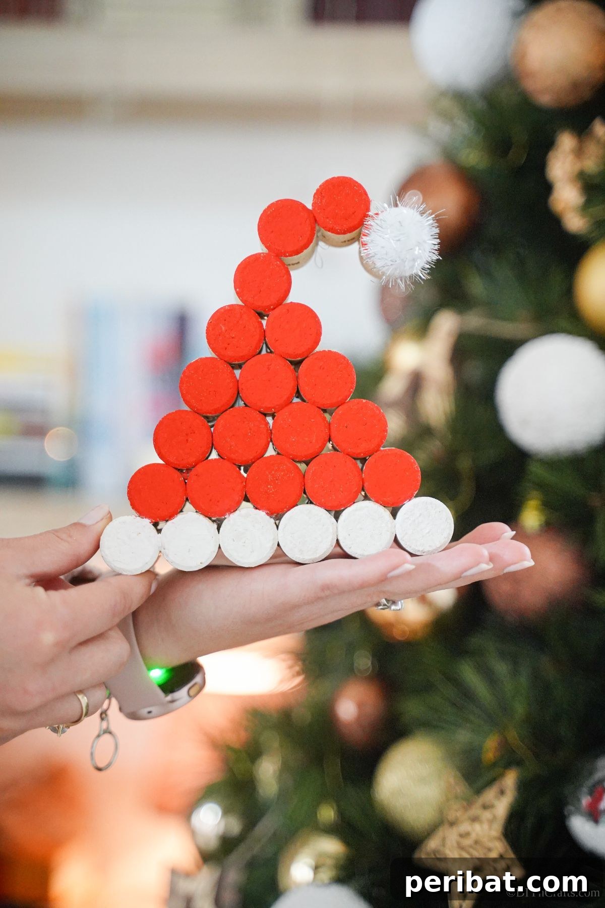 A hand lovingly holds a finished wine cork Santa hat in front of a beautifully decorated Christmas tree, showcasing the ornament's festive charm.