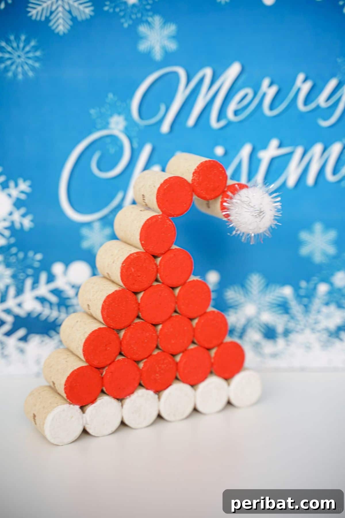 A completed wine cork Santa hat decoration rests on a white table, set against a cheerful blue and white 'Merry Christmas' sign, ready for the holiday season.
