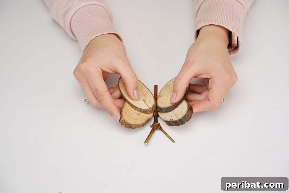 A hand applying hot glue to attach the wood slice wings to the central branch body of the butterfly, showcasing a key assembly step