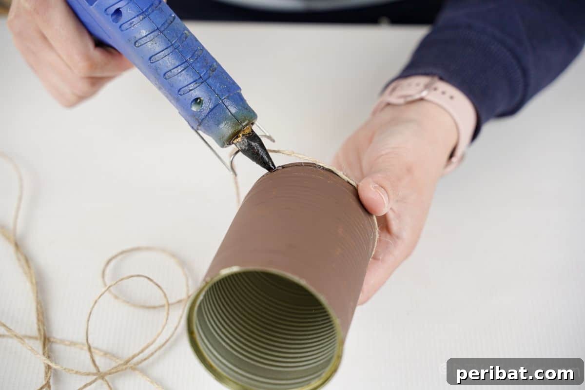 Applying hot glue and wrapping twine onto a brown-painted tin can, beginning the decoration.