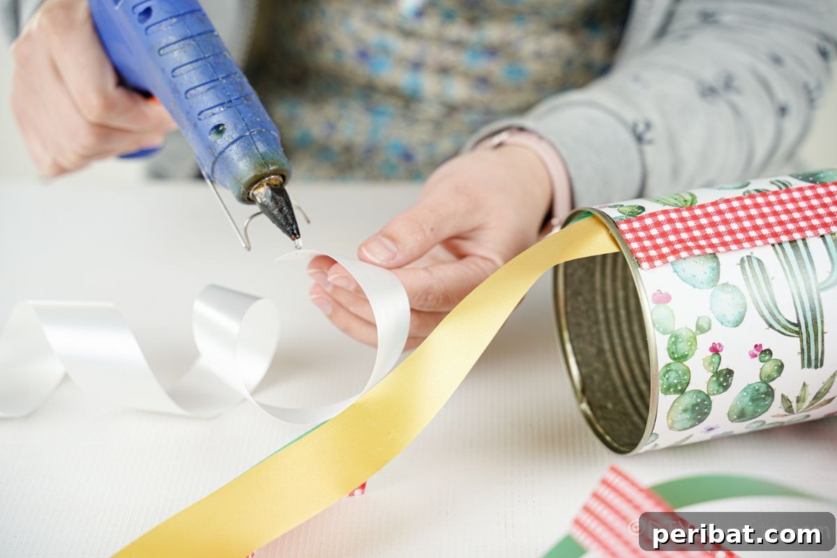 Vibrant Upcycled Tin Can Wind Socks 15 A hand carefully applying hot glue to the end of a white ribbon, preparing it to be attached inside the tin can for the windsock.