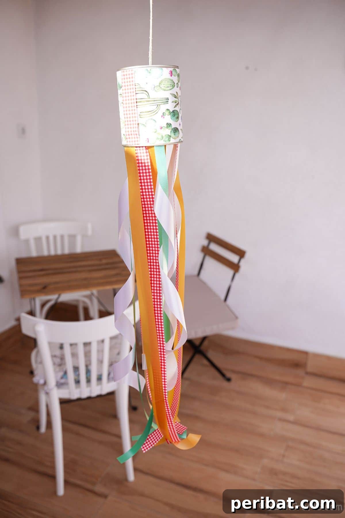 Vibrant Upcycled Tin Can Wind Socks 19 A stunning tin can windsock, with its vibrant ribbons trailing, hangs gracefully in front of a small outdoor table with white chairs, inviting peaceful enjoyment.
