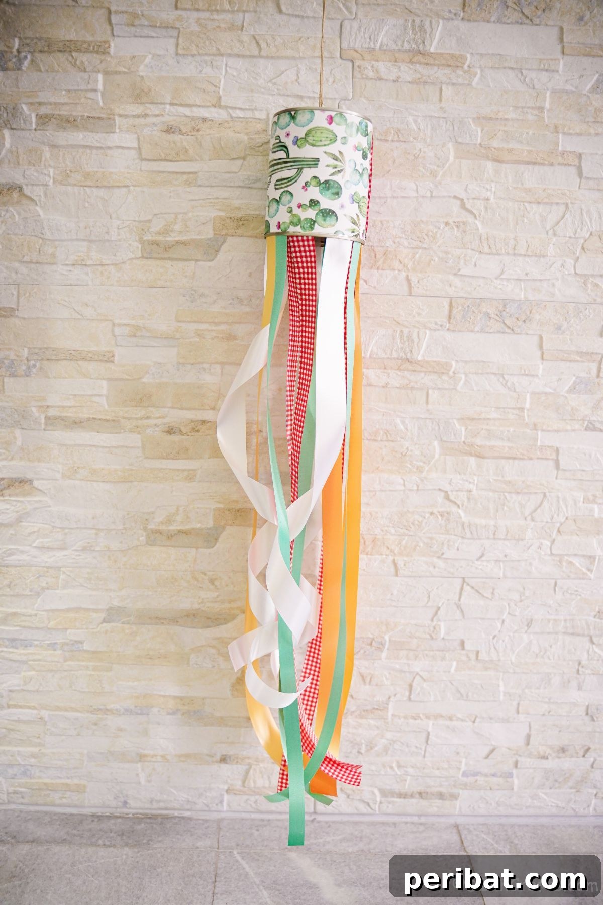 Vibrant Upcycled Tin Can Wind Socks 6 A close-up of the base of a tin can windsock, showing an assortment of colorful ribbons hanging from it, set against a rustic brick background.