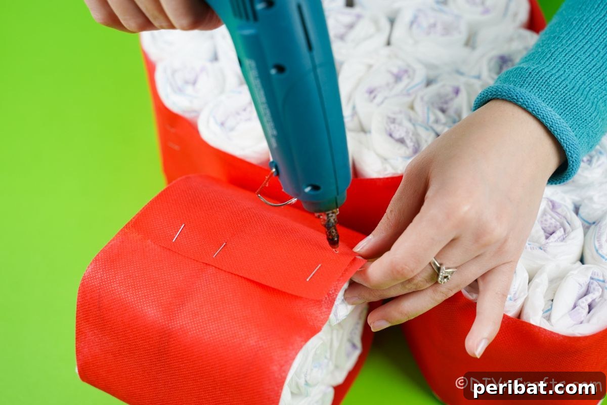 A hand using a hot glue gun to attach two smaller bundles of diapers to a larger bundle, forming Minnie's head.