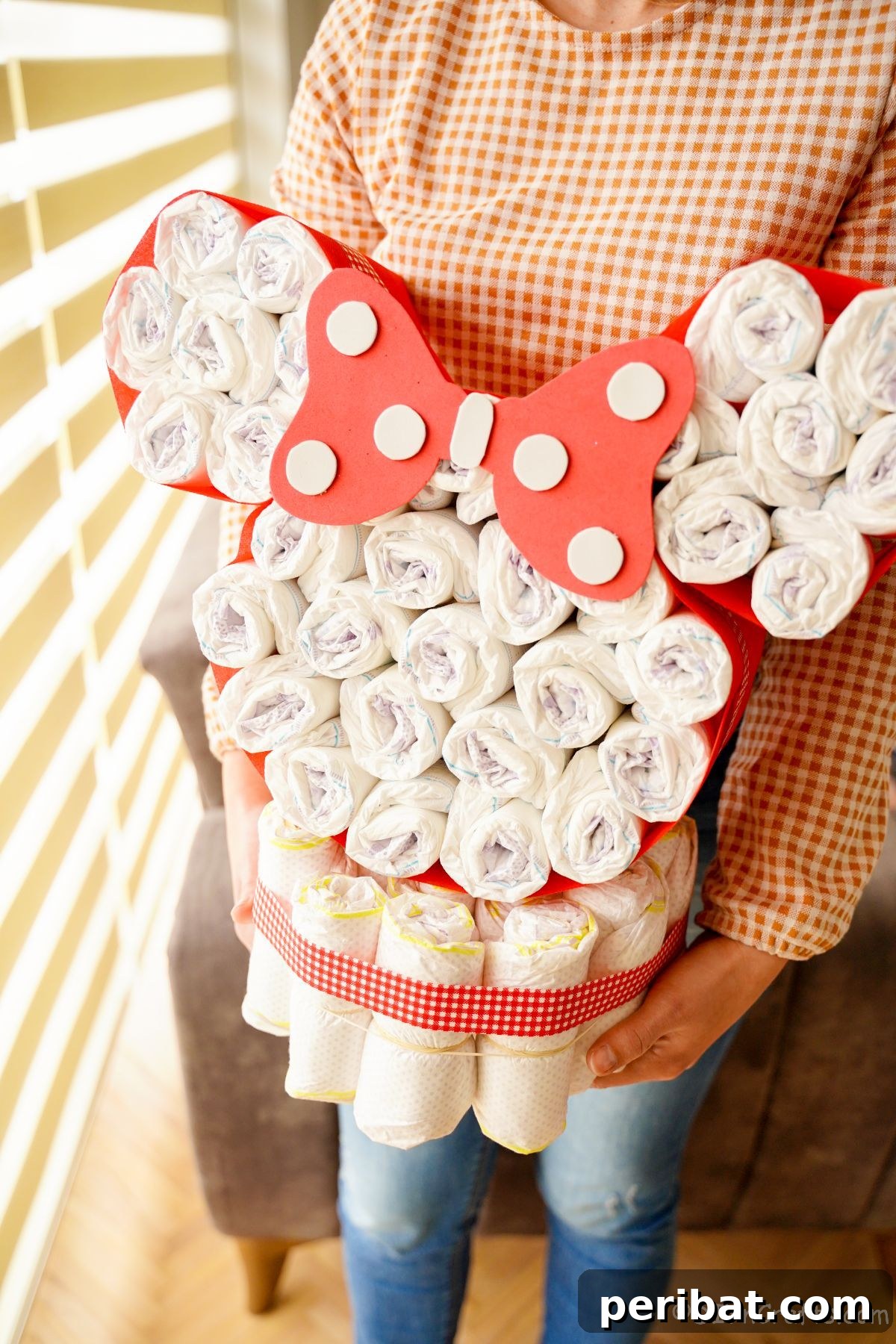 A woman in a checkered shirt and jeans proudly holds up a finished Minnie Mouse diaper cake, showcasing its size and design.