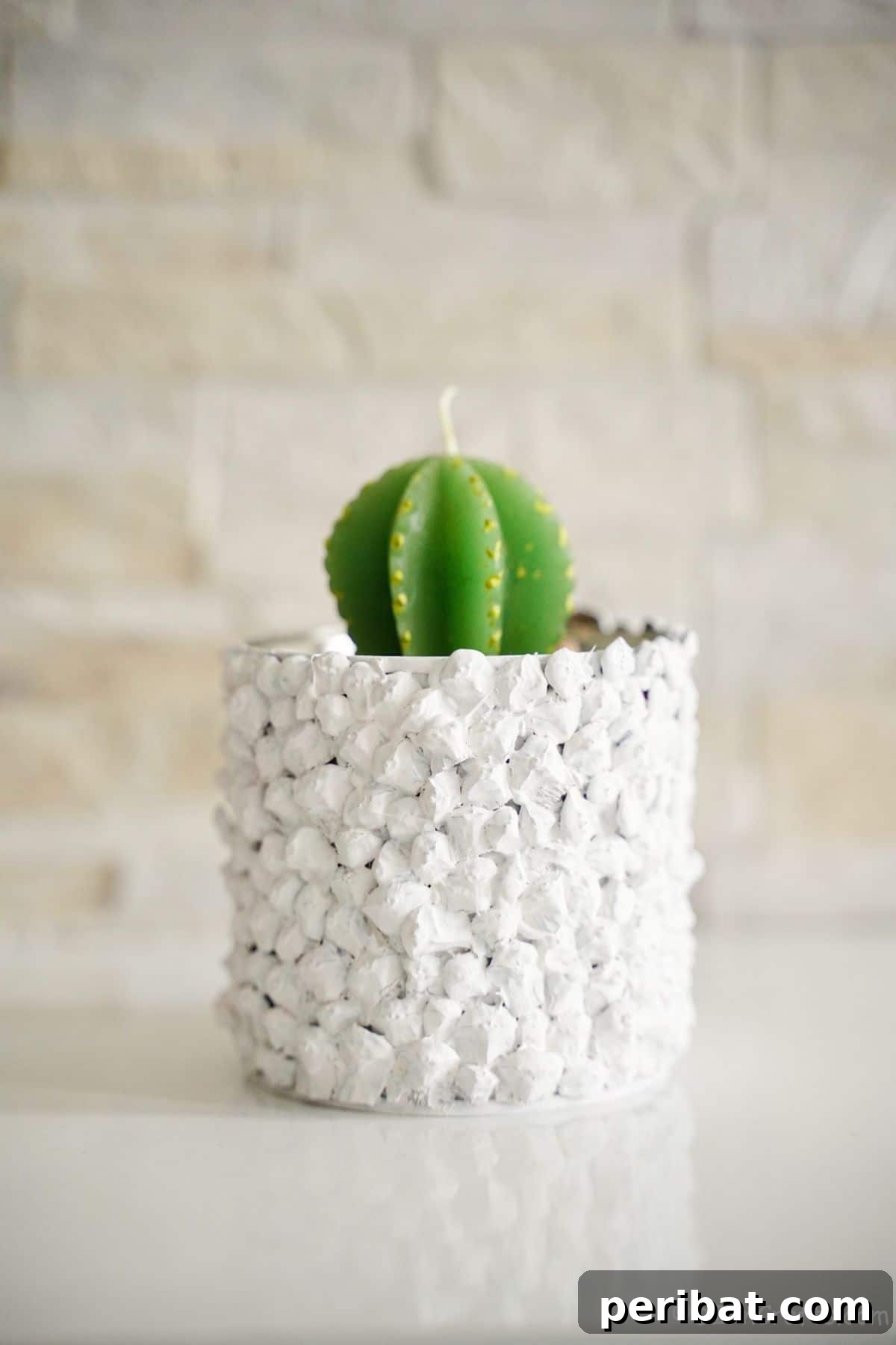 Nature-Inspired Upcycled Tin Can Planters 15 A textured white upcycled tin can planter, holding a green cactus candle, stands against a rustic brick wall, showcasing its finished beauty.