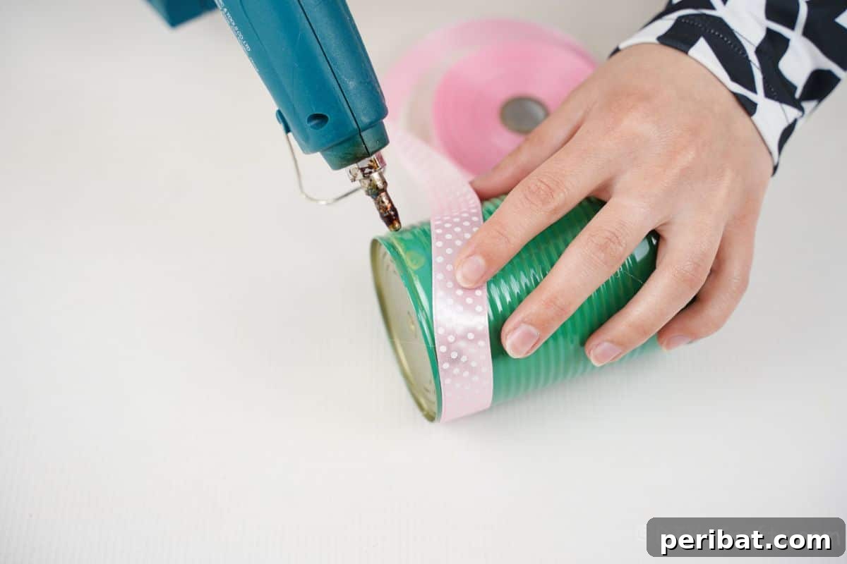 Charming Upcycled Tin Door Wreath 15 A hand is meticulously gluing a pink ribbon onto a green-painted tin can, demonstrating a step in the process of decorating for a craft project.