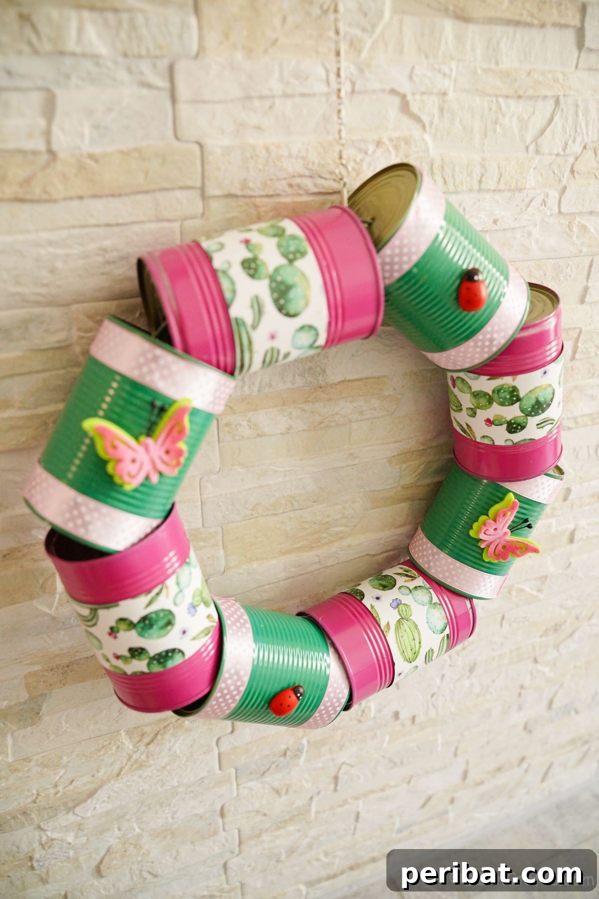 Charming Upcycled Tin Door Wreath 4 A vibrant tin can wreath, painted in pink and green, hangs against a rustic cream brick wall, adding a cheerful splash of color to the outdoor space.