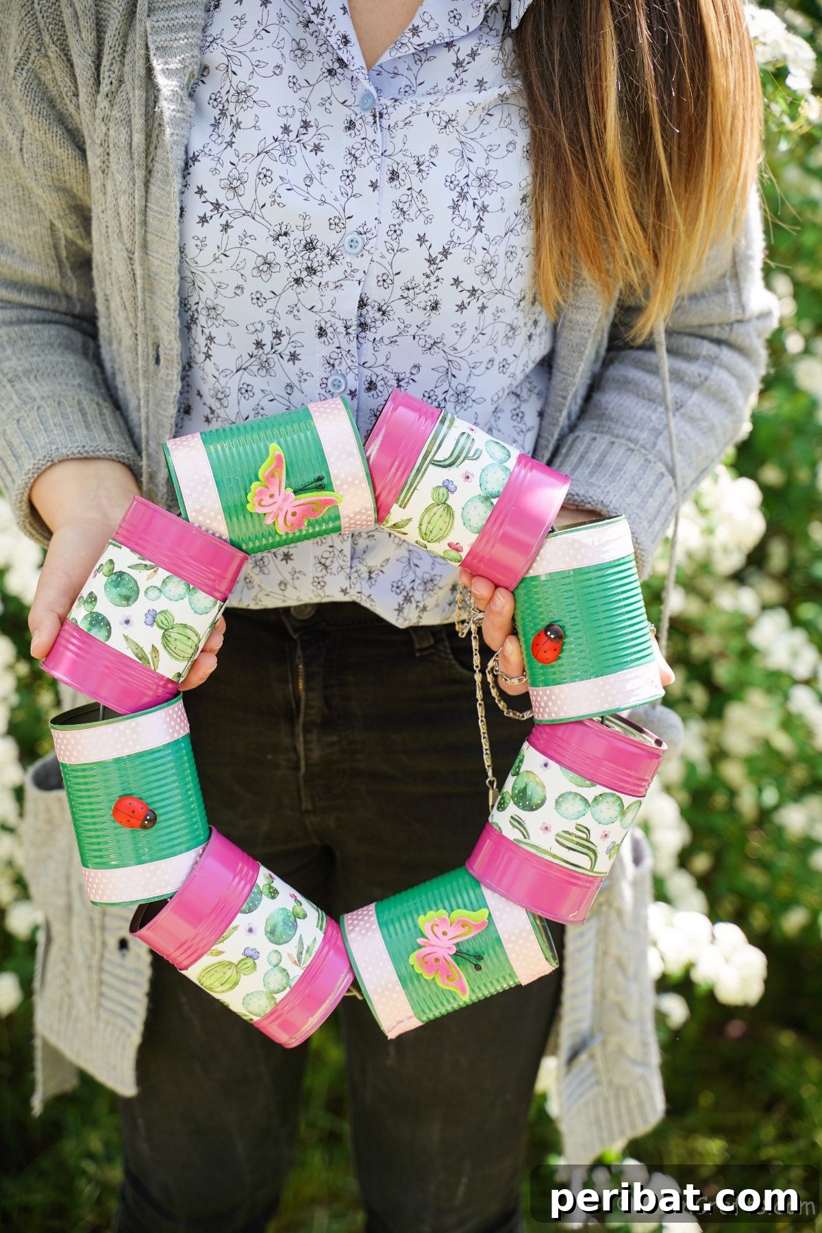 Charming Upcycled Tin Door Wreath 6 A woman in a light-colored top gently holds a vibrant pink and green tin can wreath, with lush green shrubs providing a soft background.
