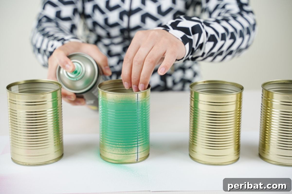 Charming Upcycled Tin Door Wreath 9 A hand is actively spraying green paint onto several empty tin cans arranged on a protective surface, preparing them for a craft project.