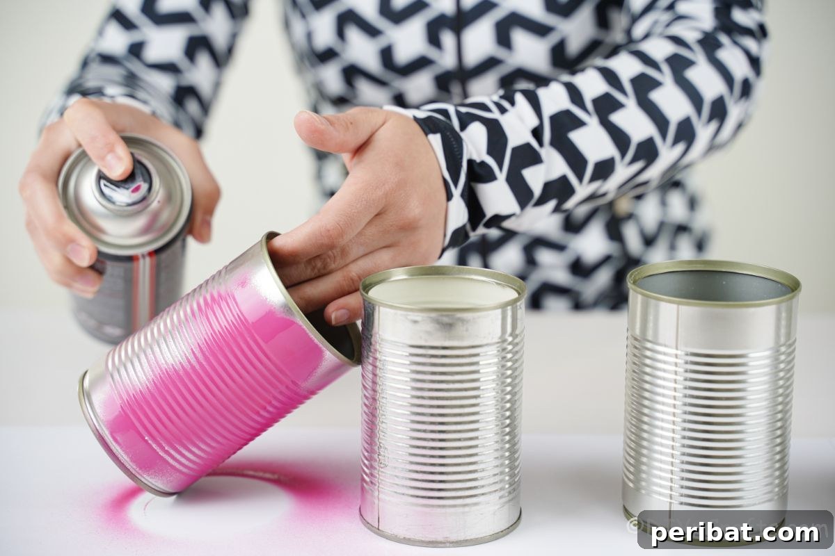 Charming Upcycled Tin Door Wreath 10 A hand applies pink spray paint to a cluster of tin cans, preparing them for a colorful craft project, set on a protected surface.