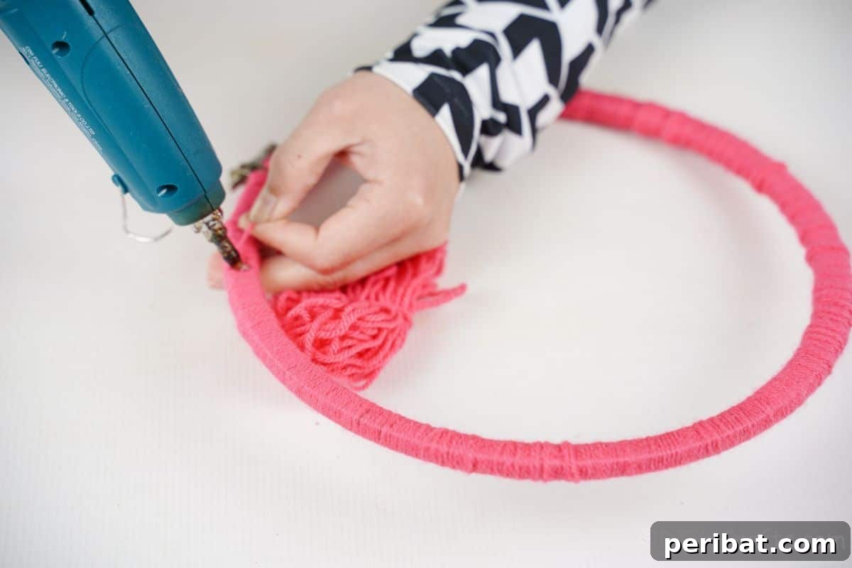 Transform Tin Cans into Melodious Wind Chimes 11 Vibrant pink yarn expertly wrapped around an embroidery hoop, with a hand applying hot glue to secure the wrapping in place