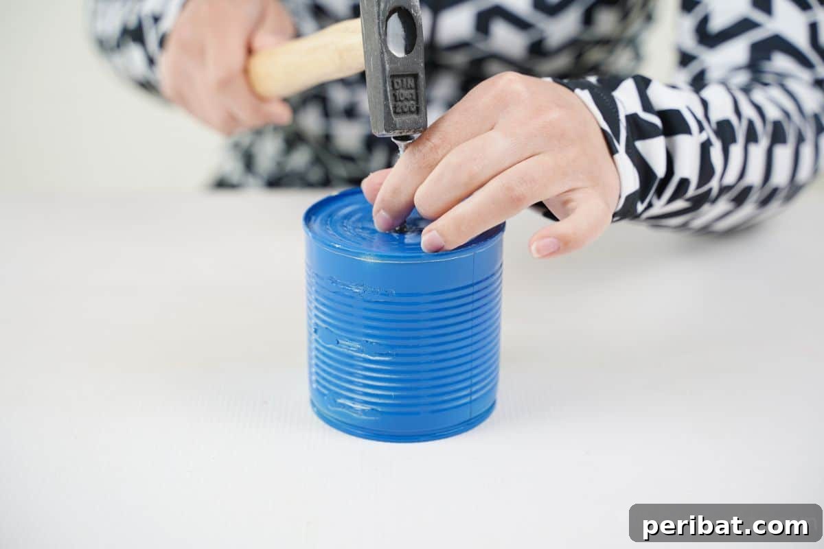 Transform Tin Cans into Melodious Wind Chimes 14 A hammer and a small nail are carefully positioned above a blue tin can, poised to create a precise hanging hole in its top