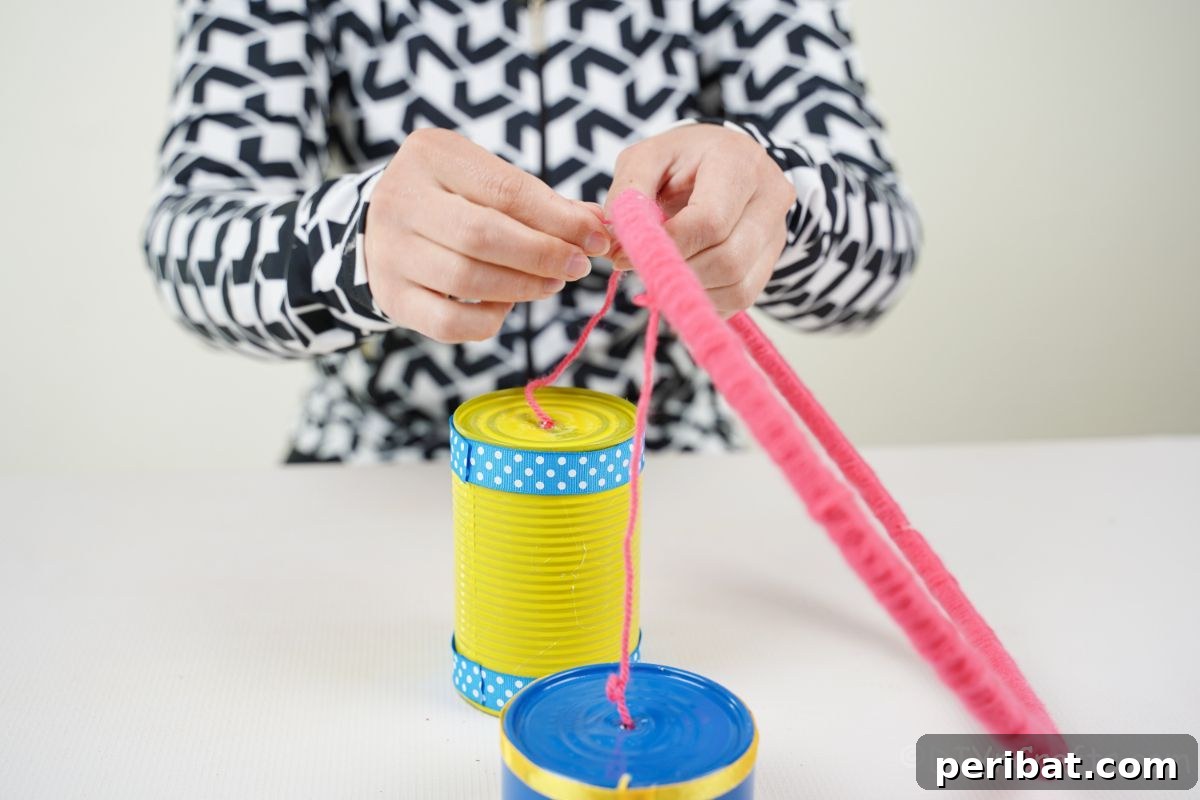 Transform Tin Cans into Melodious Wind Chimes 16 A hand carefully tying a vibrant tin can, suspended by pink yarn, to a perfectly pink yarn-wrapped embroidery hoop, showcasing the assembly process