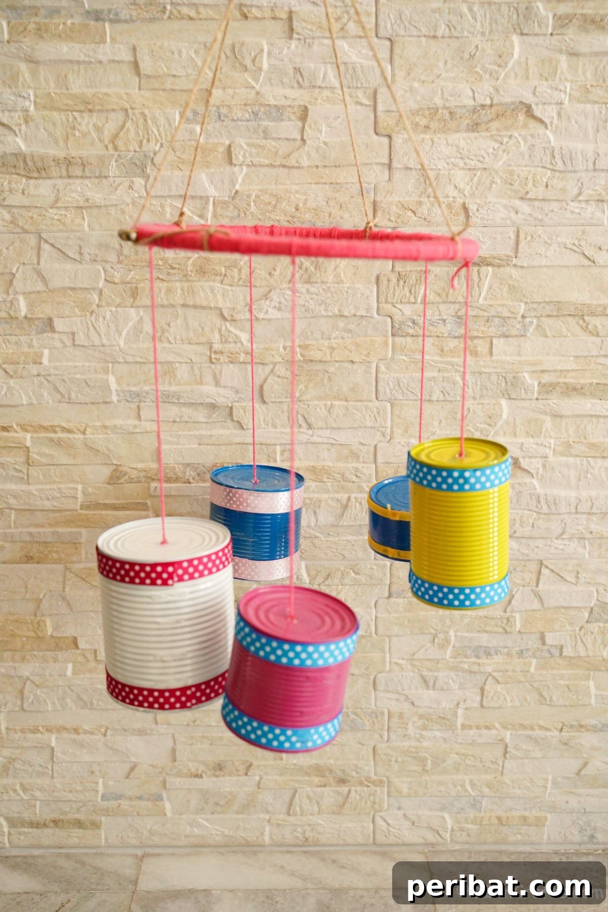 Transform Tin Cans into Melodious Wind Chimes 6 An elegant tin can wind chime with a delicate pink base suspended gracefully against a rustic, textured cream brick wall