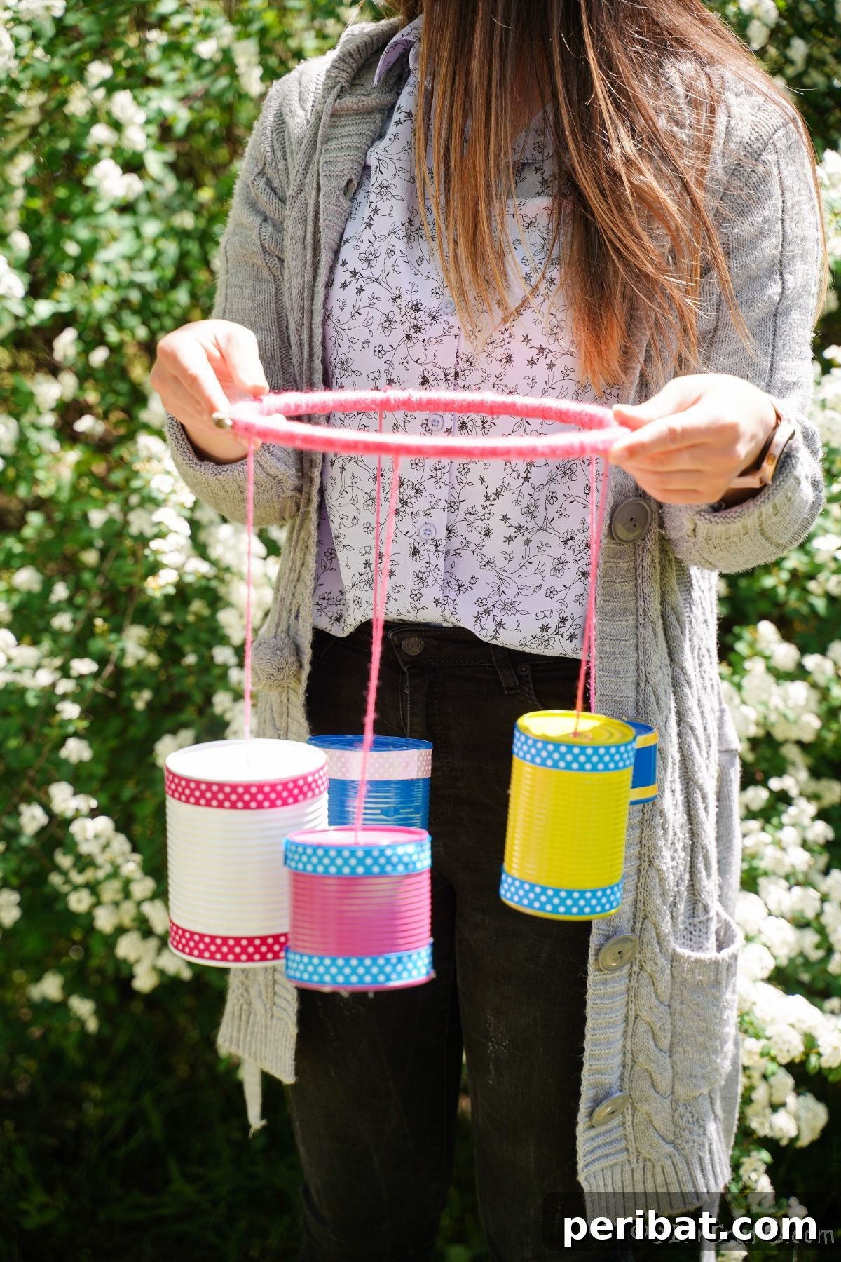 Transform Tin Cans into Melodious Wind Chimes 7 A creative woman proudly presenting her vibrant, handmade tin can wind chime against a serene backdrop of white shrubs, highlighting its beauty