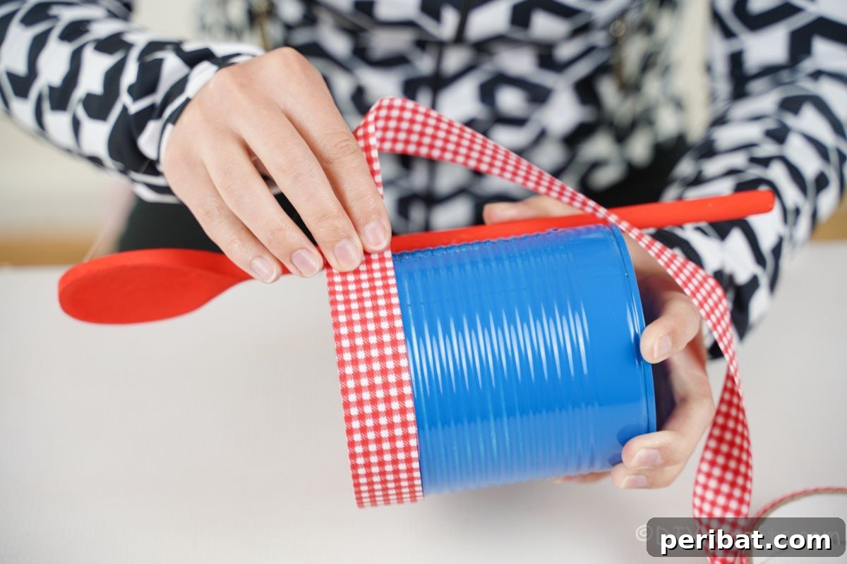 Sweet DIY Tin Can Bird Feeder 11 A hand neatly wraps red and white checked ribbon over the edge of a blue tin can, adding a decorative and securing element.