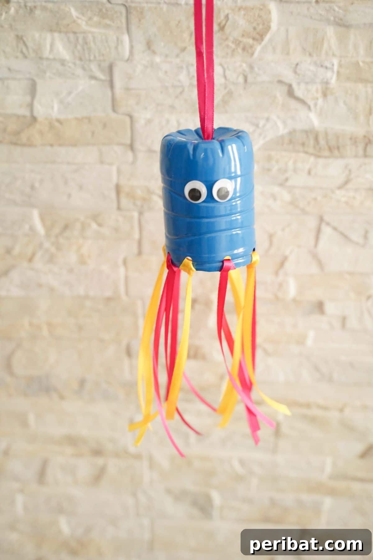 Vibrant Upcycled Bottle Wind Streamer 5 Blue, pink, and yellow plastic bottle windsock against a cream brick wall