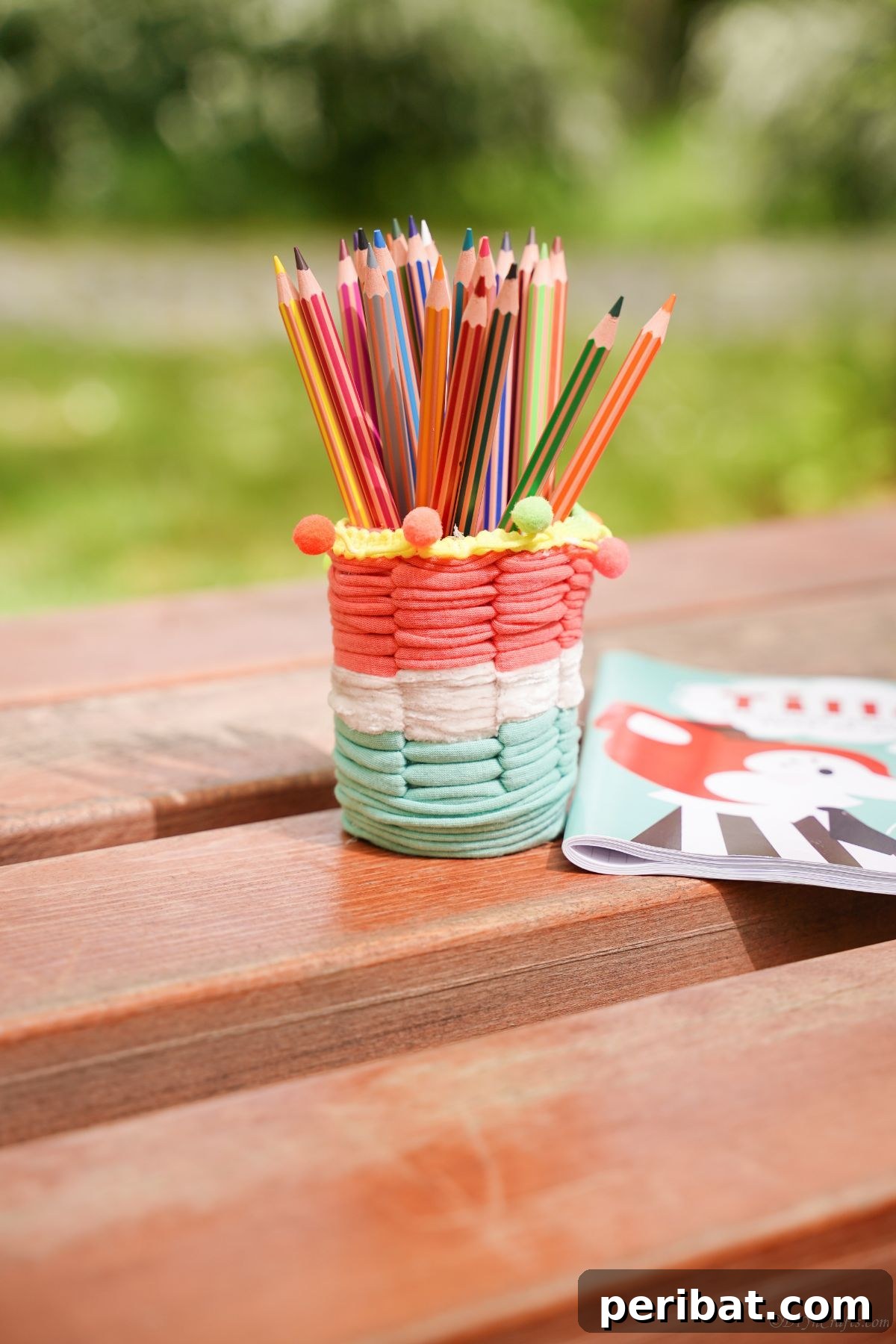 Upcycled Plastic Bottle Desk Organizer 3 Colorful striped pencil cup, crafted from a plastic bottle and covered in white, green, and pink yarn, resting on a picnic table.