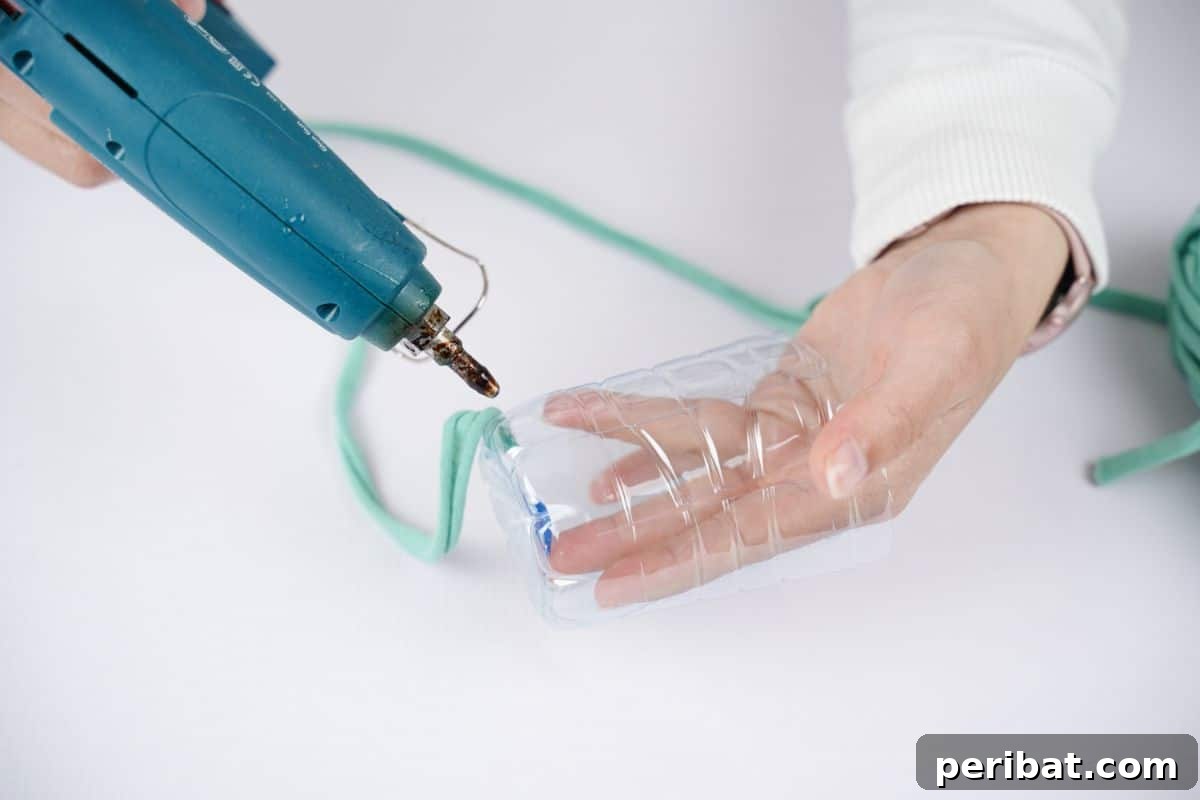 Upcycled Plastic Bottle Desk Organizer 10 A blue hot glue gun expertly attaching vibrant green yarn to the bottom edge of a plastic bottle, initiating the weaving process.