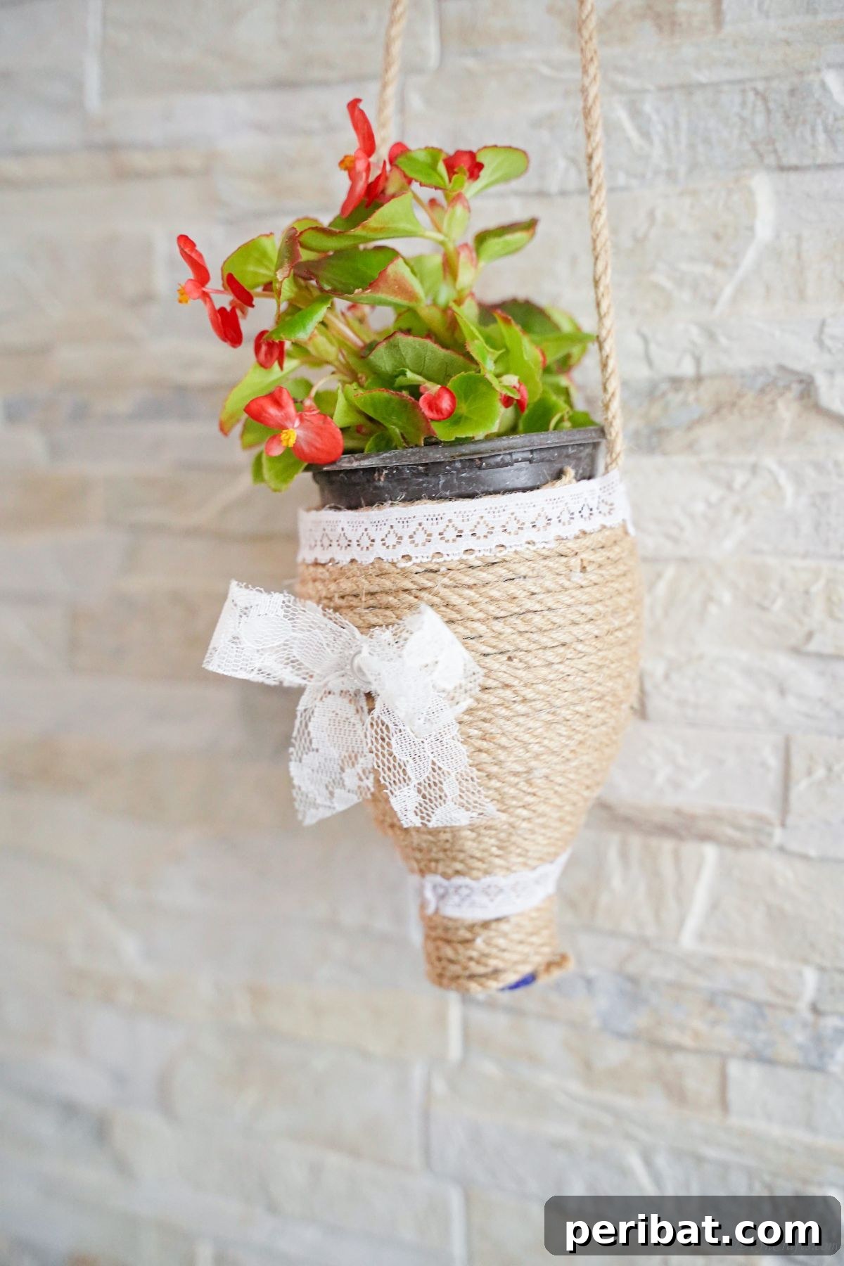DIY Rustik İp Sarılı Şişe Bahçe Planterları 6 A beautiful twine planter holding vibrant red flowers, hanging gracefully against a cream brick wall, enhancing outdoor aesthetic.