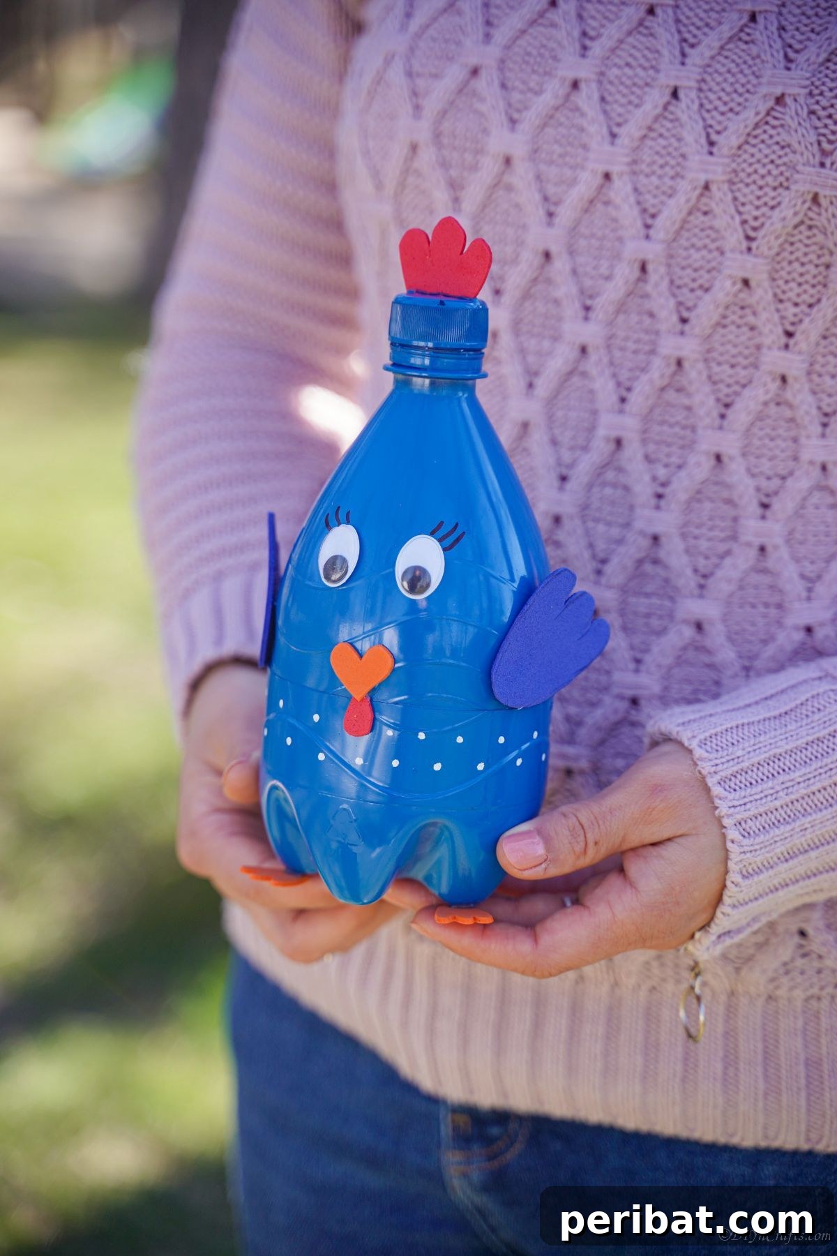 A woman in a pink sweater holding the finished blue plastic bottle bird craft.