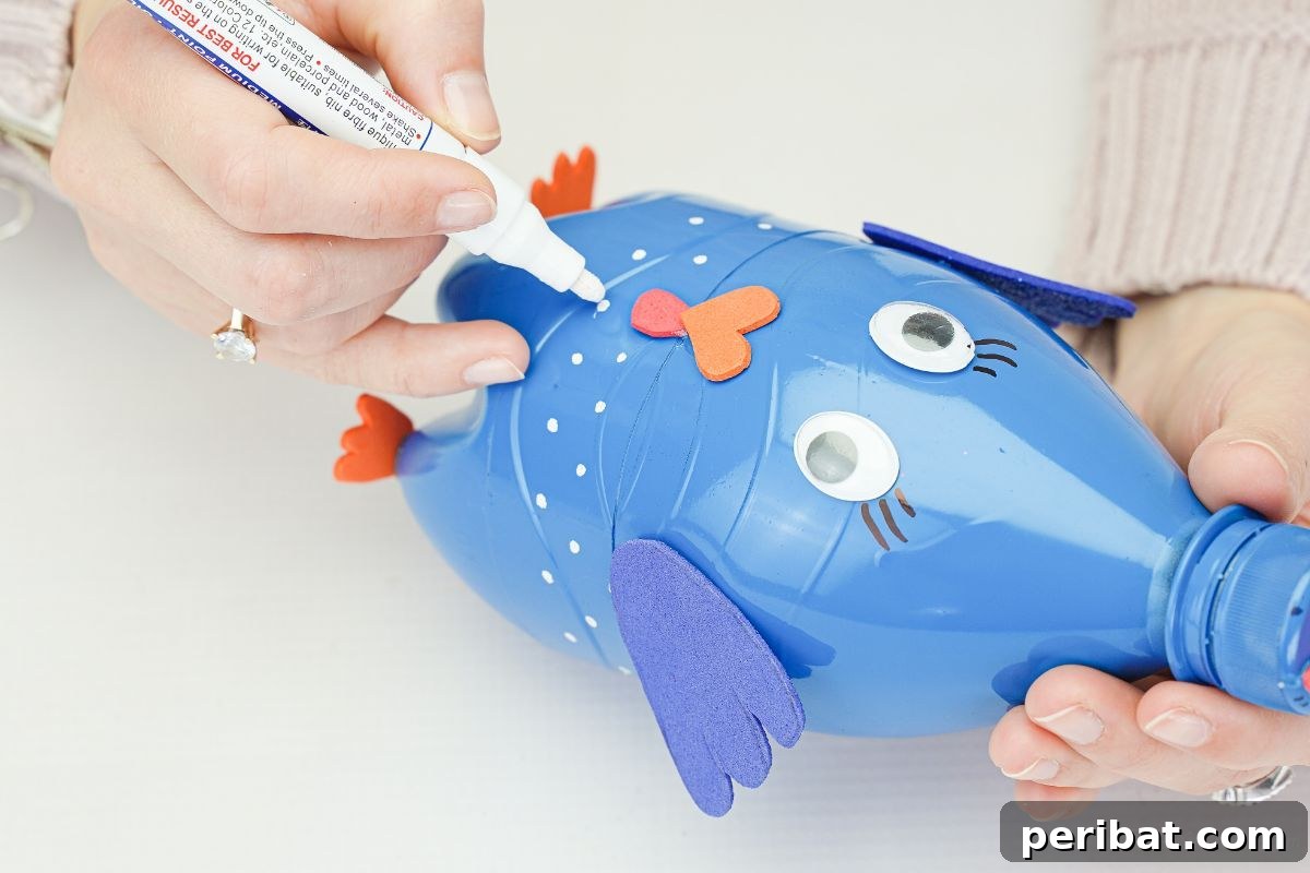 Hand using a white paint pen to make decorative dots on the blue plastic bottle turkey.