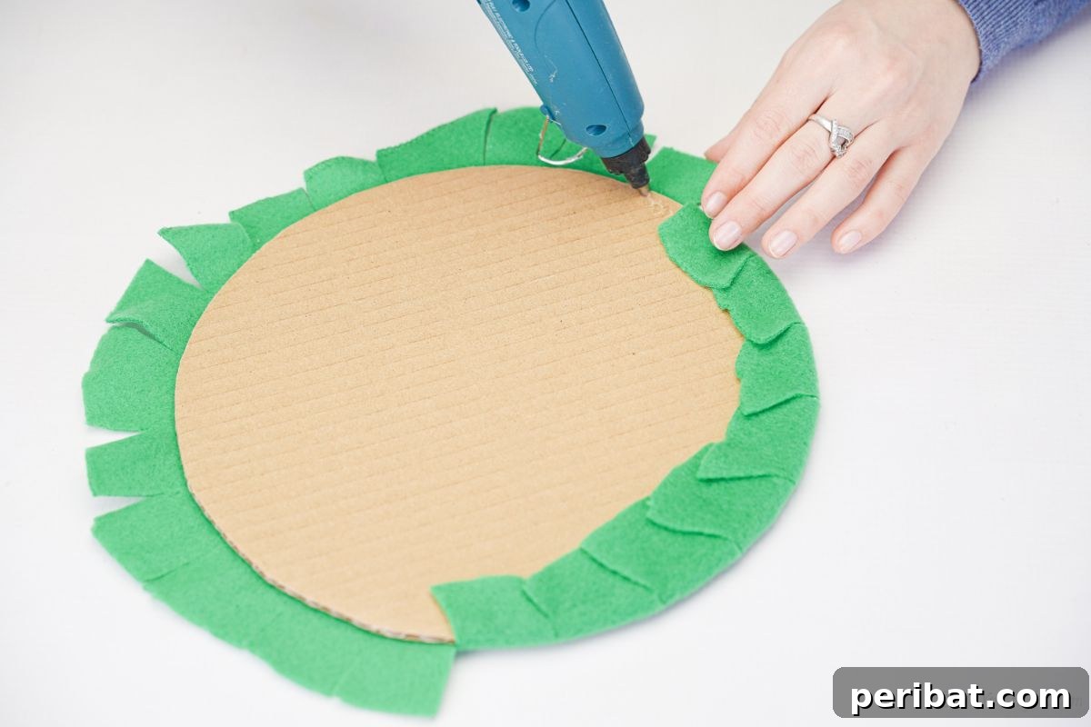Hand using a hot glue gun to attach green felt over the edge of the cardboard circle, showing the neat finish.