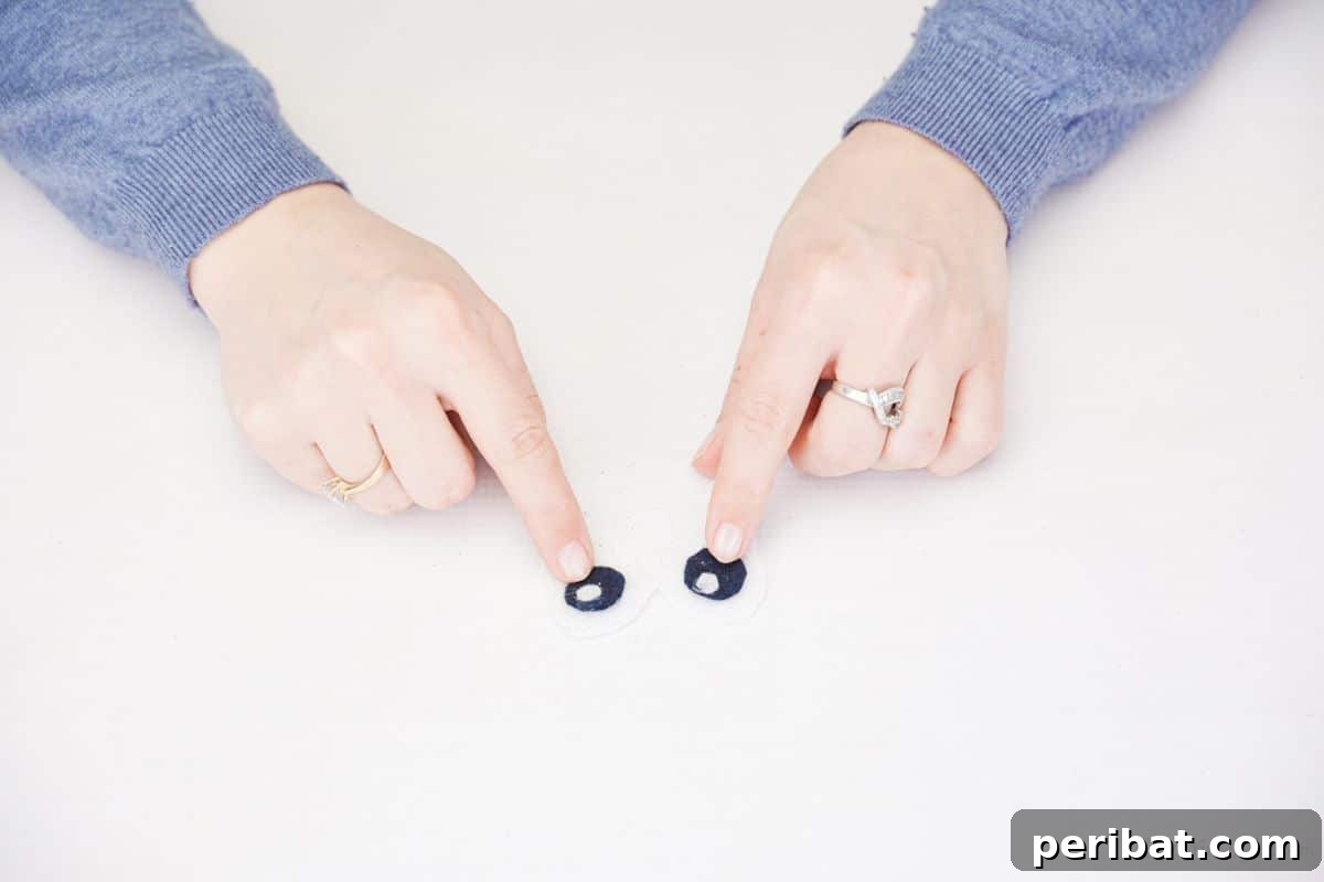 Fingers holding two black felt eyes with small white highlights, positioned on the white heart-shaped dog face.