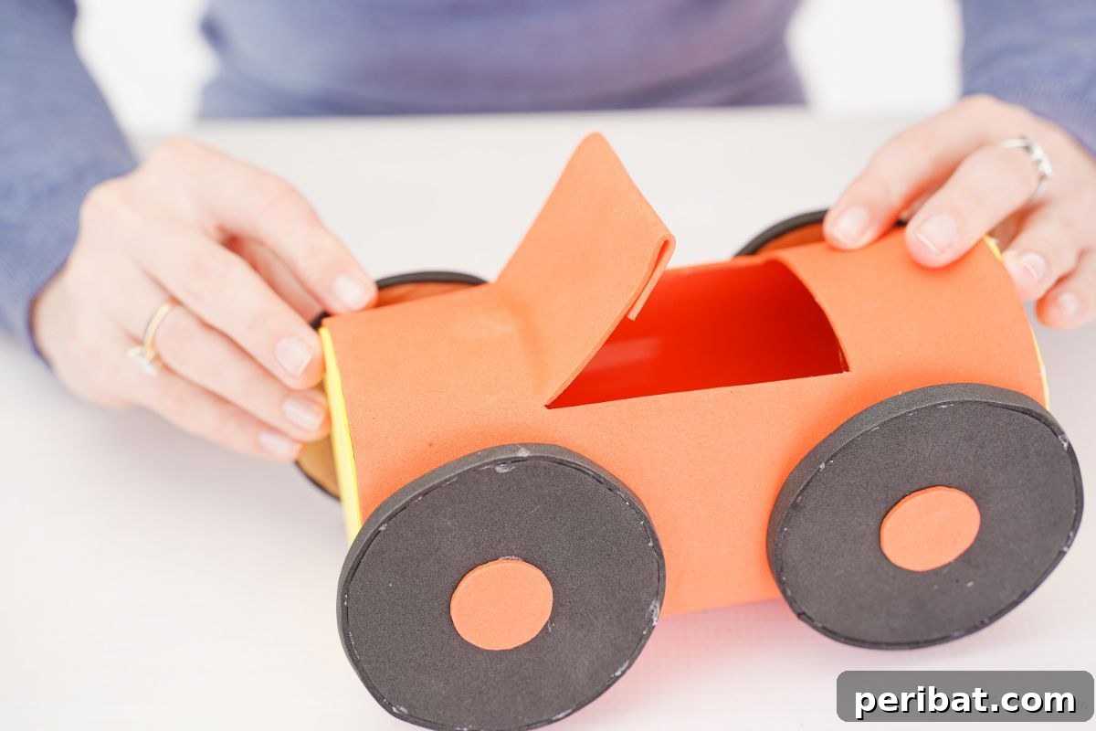 A hand holding two black and orange wheels against the side of the orange plastic bottle car body, ready for gluing.