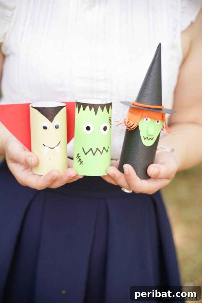 A woman holding a charming trio of Halloween characters crafted from toilet paper rolls: a witch, a vampire, and Frankenstein's monster.