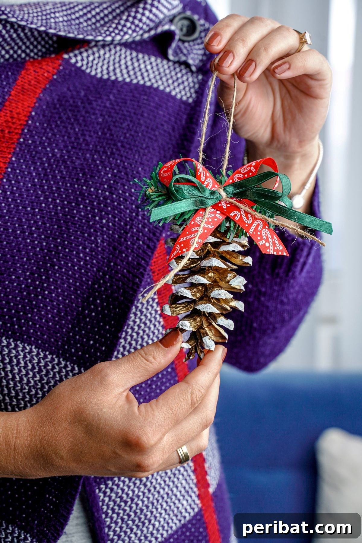 DIY Pinecone Christmas Ornaments 14 A woman in a purple sweater holding a finished pinecone ornament with white frosted tips and a red ribbon