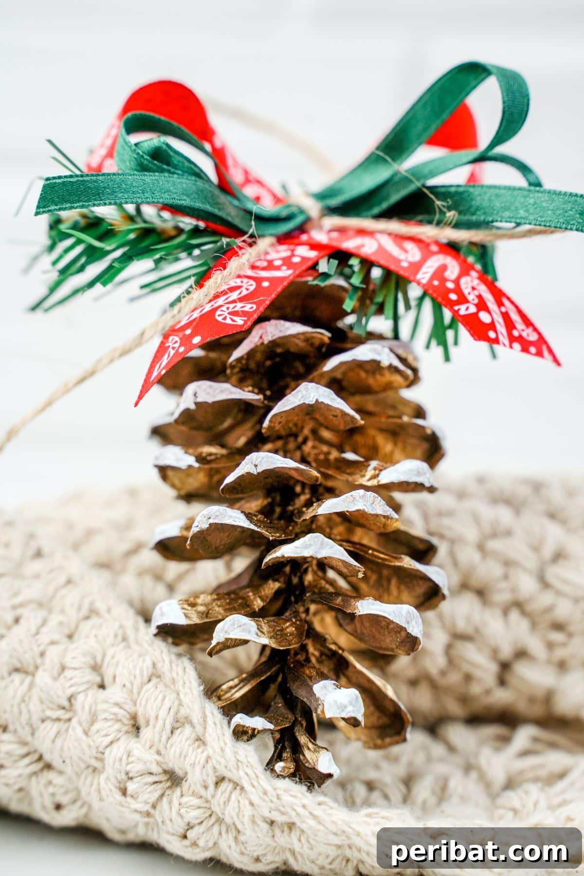 DIY Pinecone Christmas Ornaments 16 A beautifully crafted pinecone ornament with white frosted tips and a red ribbon, resting on a soft cream blanket