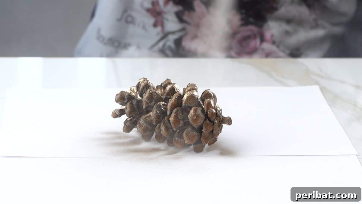 DIY Pinecone Christmas Ornaments 5 Close-up of a hand spray painting a pinecone with gold paint on a protective surface
