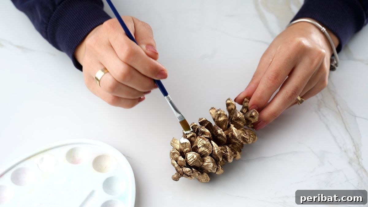 DIY Pinecone Christmas Ornaments 6 A hand gently applying white paint to the tips of a pinecone's scales, creating a frosted effect