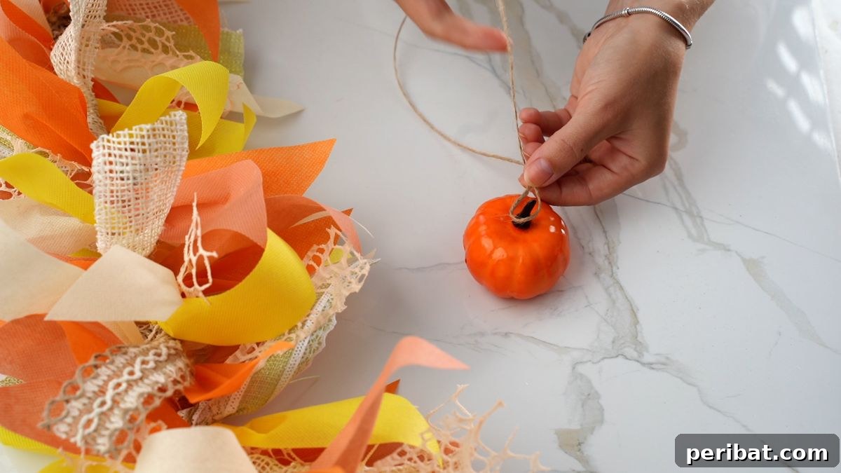 A hand tying twine around the stem of a small fake pumpkin to attach it to the garland