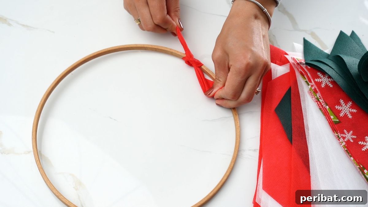 Effortless Festive Fabric Wreath 7 A hand carefully tying a vibrant red fabric strip onto the bare embroidery hoop, forming the base of the Christmas rag wreath.