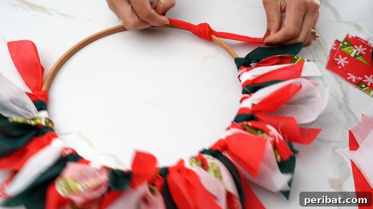 Effortless Festive Fabric Wreath 9 A hand securing another red fabric strip onto the wreath form, gradually building up the volume and color of the rag wreath.