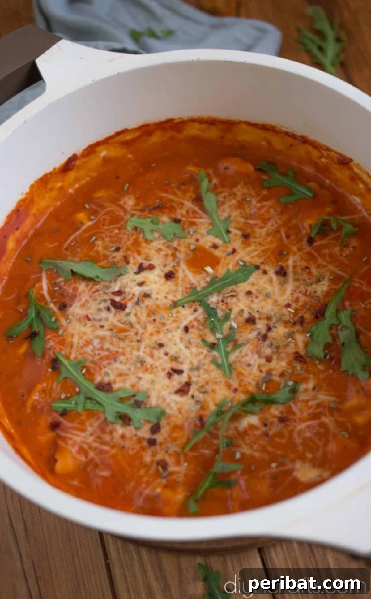 A hand garnishing a bowl of tomato tortellini soup with fresh green basil leaves and grated parmesan cheese.