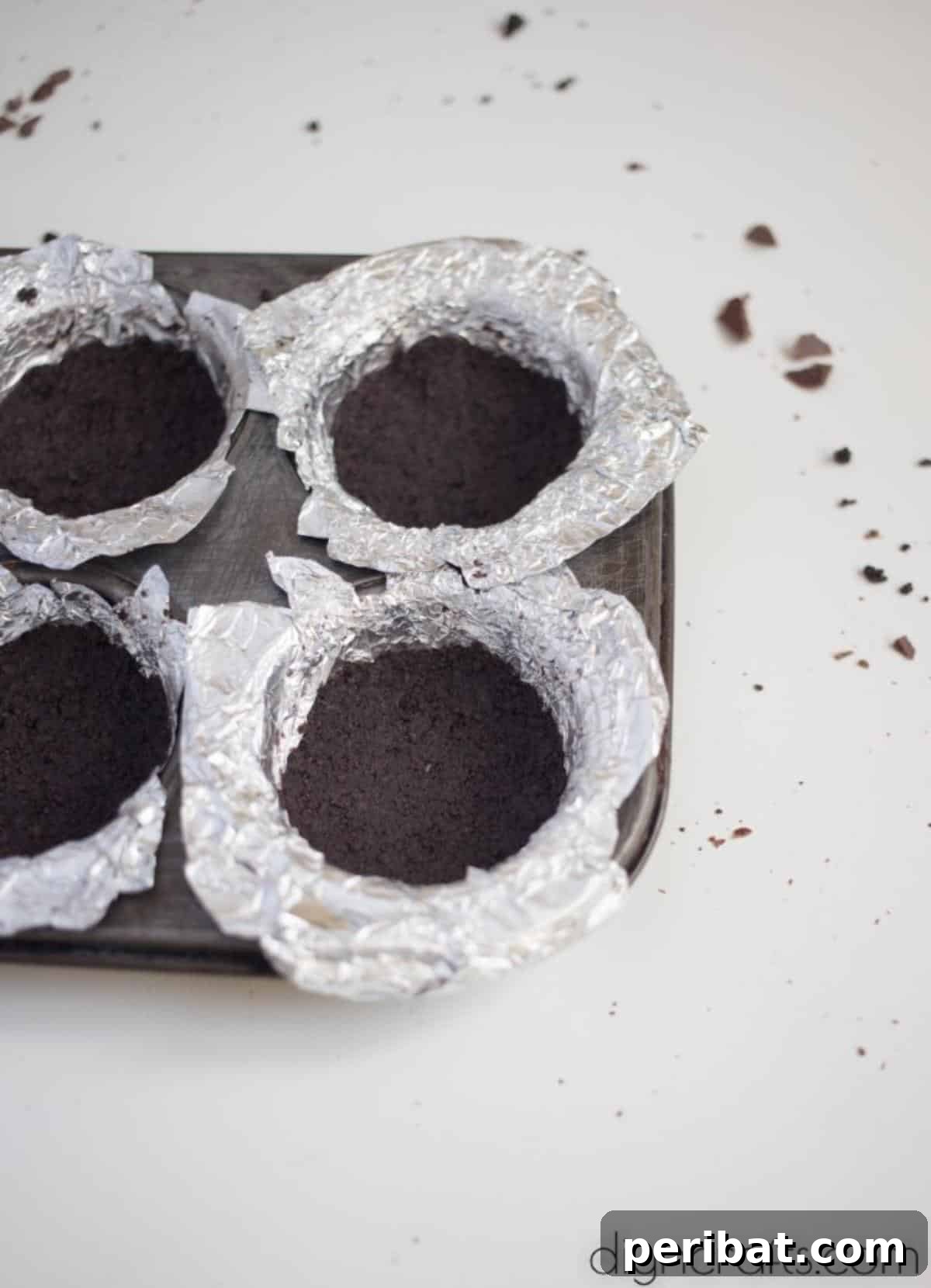 Crush cookies, mix with butter, and create crusts in lined cupcake pans - A hand pressing a mixture of crushed Oreos and melted butter into a foil-lined cupcake mold to form the crust.
