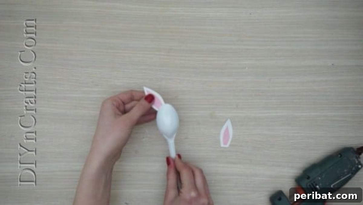 Spoonfuls of Fun 5 Easter Crafts for Kids Using Plastic Spoons 4 A hand is carefully gluing a pair of white and pink paper bunny ears onto the back of a plastic spoon.