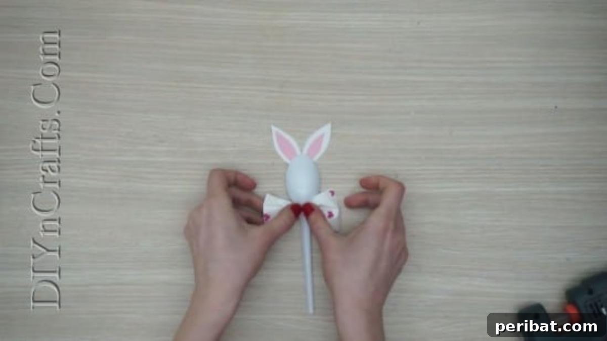 Spoonfuls of Fun 5 Easter Crafts for Kids Using Plastic Spoons 6 A small pink decorative bow is being glued onto the handle of the plastic spoon, just below the bunny's head, to create a bow tie.