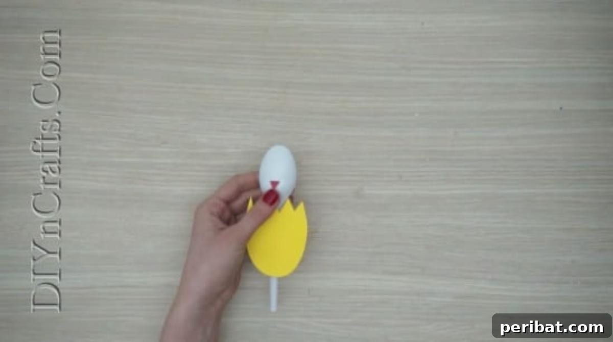 Spoonfuls of Fun 5 Easter Crafts for Kids Using Plastic Spoons 10 A small orange paper triangle is being glued onto the plastic spoon to form the chick's beak, positioned above the yellow eggshell.