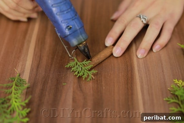 Attaching the first pine sprig to the cinnamon stick with hot glue, forming the tree base.